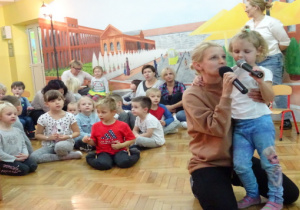 Dziewczynka z mamą śpiewa piosenkę, z tyłu siedzą rodzice z dziećmi.